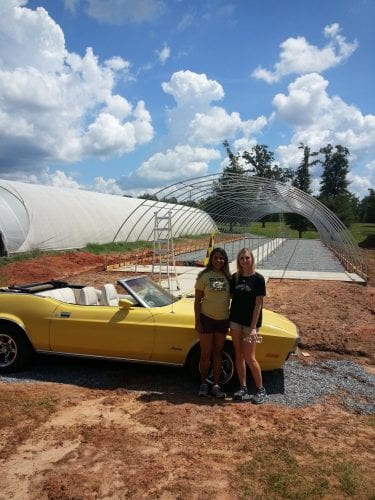 Heather Johnston poses with a friend on a farm, both standing in front of a yellow convertible with long farming houses in the background.