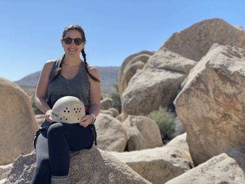 Sarah Banks Hogg poses in front of a large group of rocks while holding a helmet in her hands.