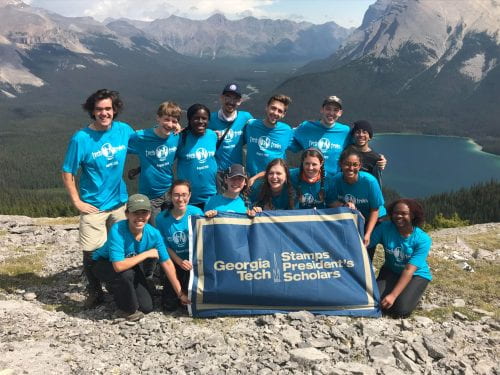 A group of students and staff stand on a mountain top all wearing turquoise 'Tech Trek' shirts. They are holding a flag that reads 'Georgia Tech Stamps President's Scholars'